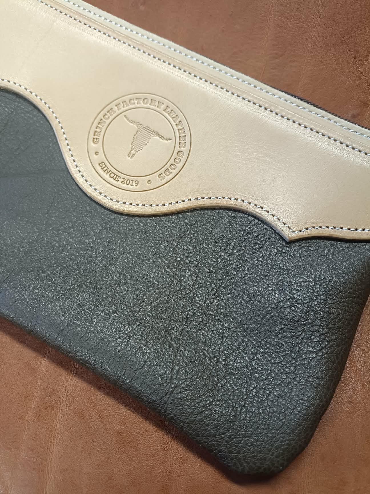 Pochette en cuir bicolore Grinch Factory Leather Goods avec logo embossé et dragonne de poignet.