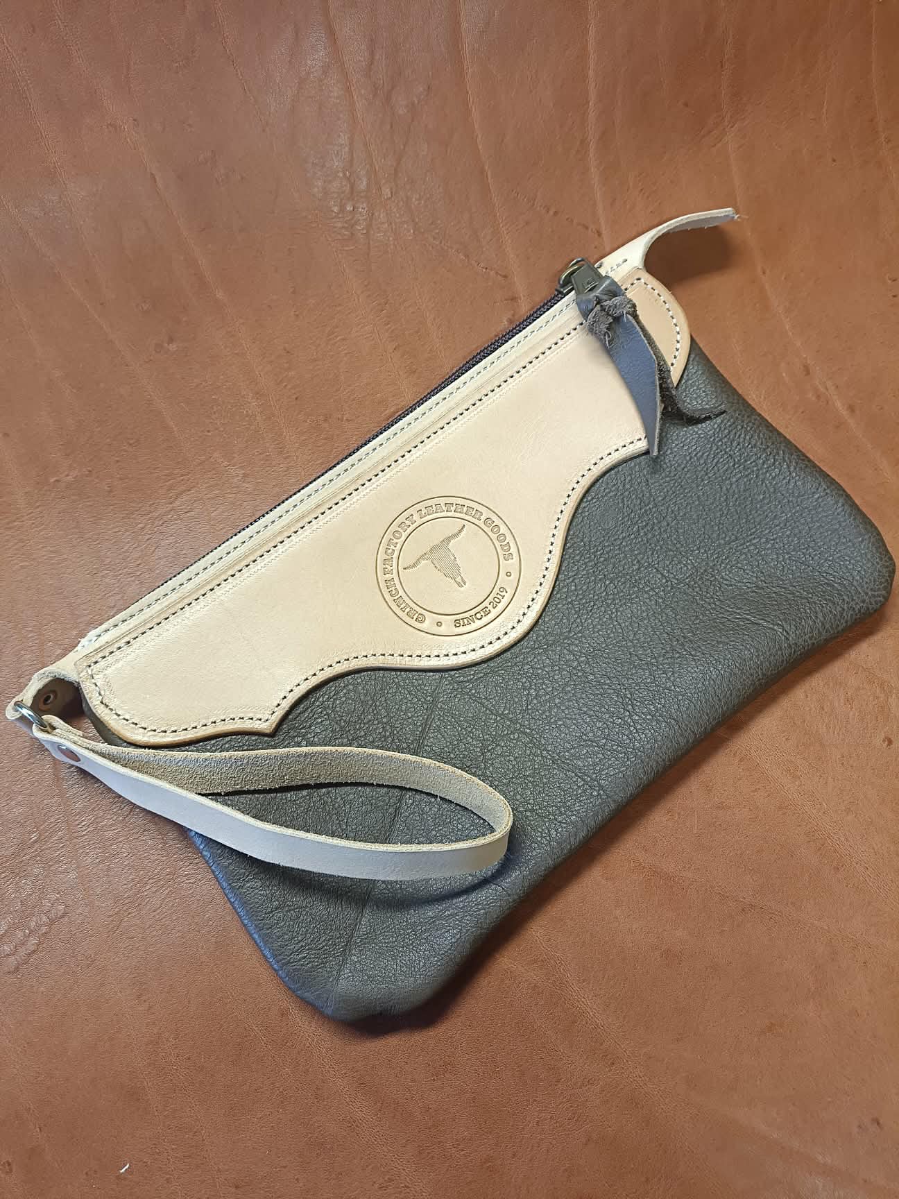 Pochette en cuir bicolore Grinch Factory Leather Goods avec logo embossé et dragonne de poignet.