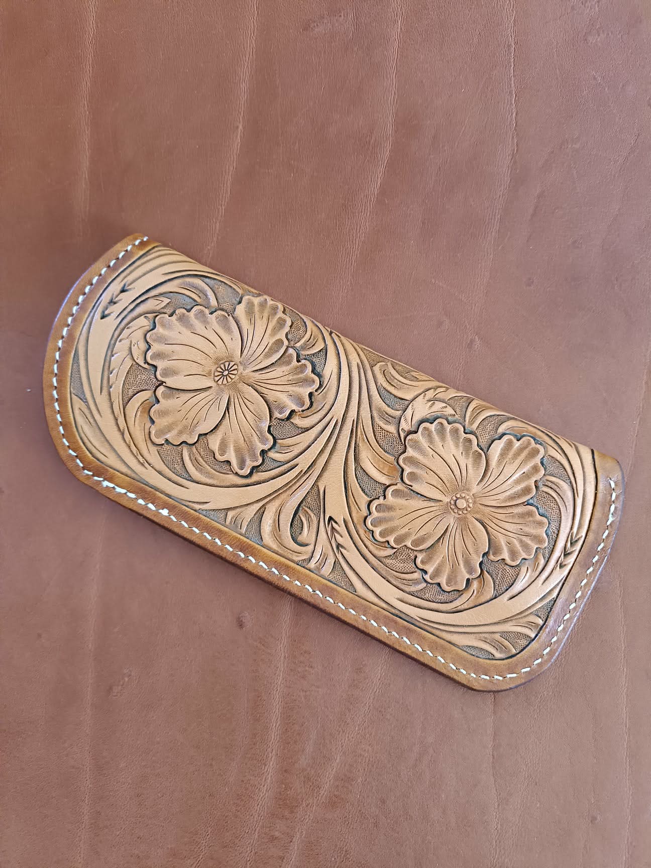 Étui à lunettes en cuir naturel avec un grand motif de fleurs sculptées en relief et coutures blanches, posé à côté d'une paire de lunettes sur un fond en cuir marron.