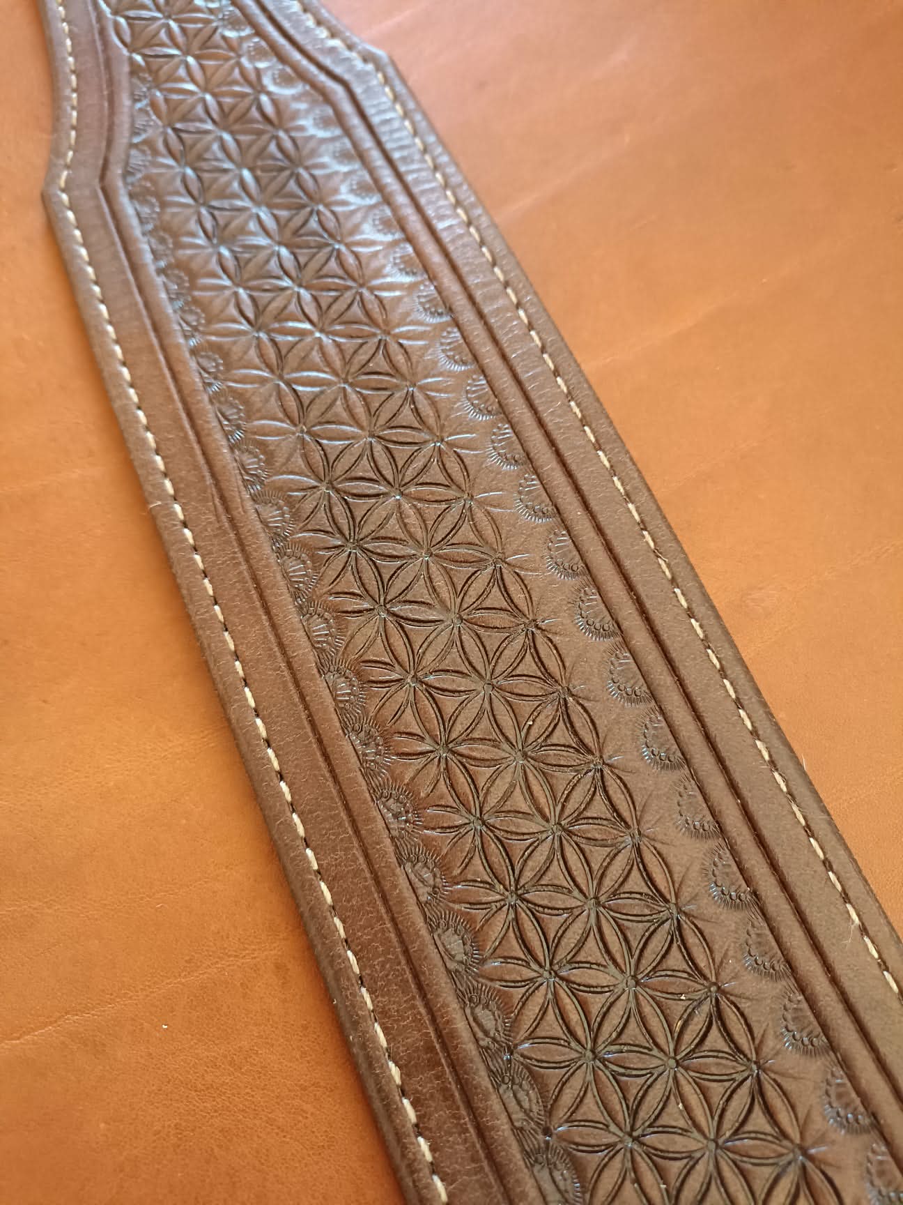 Sangle de guitare en cuir marron clair avec motif fleur de vie, épaulière large et coutures blanches sur fond de cuir naturel.