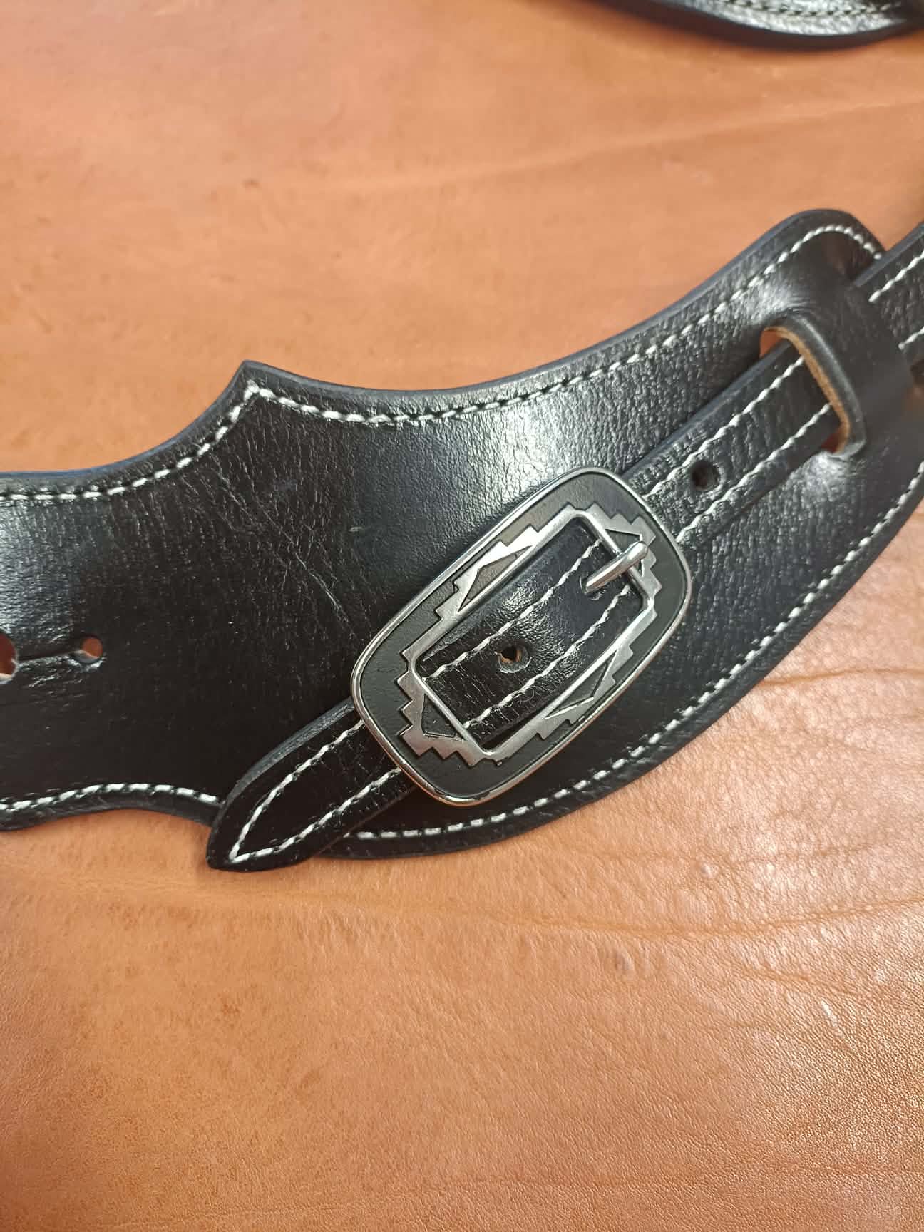 Paire de pattes d'éperons en cuir noir artisanal avec coutures blanches et boucles métalliques gravées style Western posées sur un fond en cuir marron.