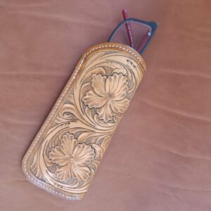Étui à lunettes en cuir naturel avec un grand motif de fleurs sculptées en relief et coutures blanches, posé à côté d'une paire de lunettes sur un fond en cuir marron.