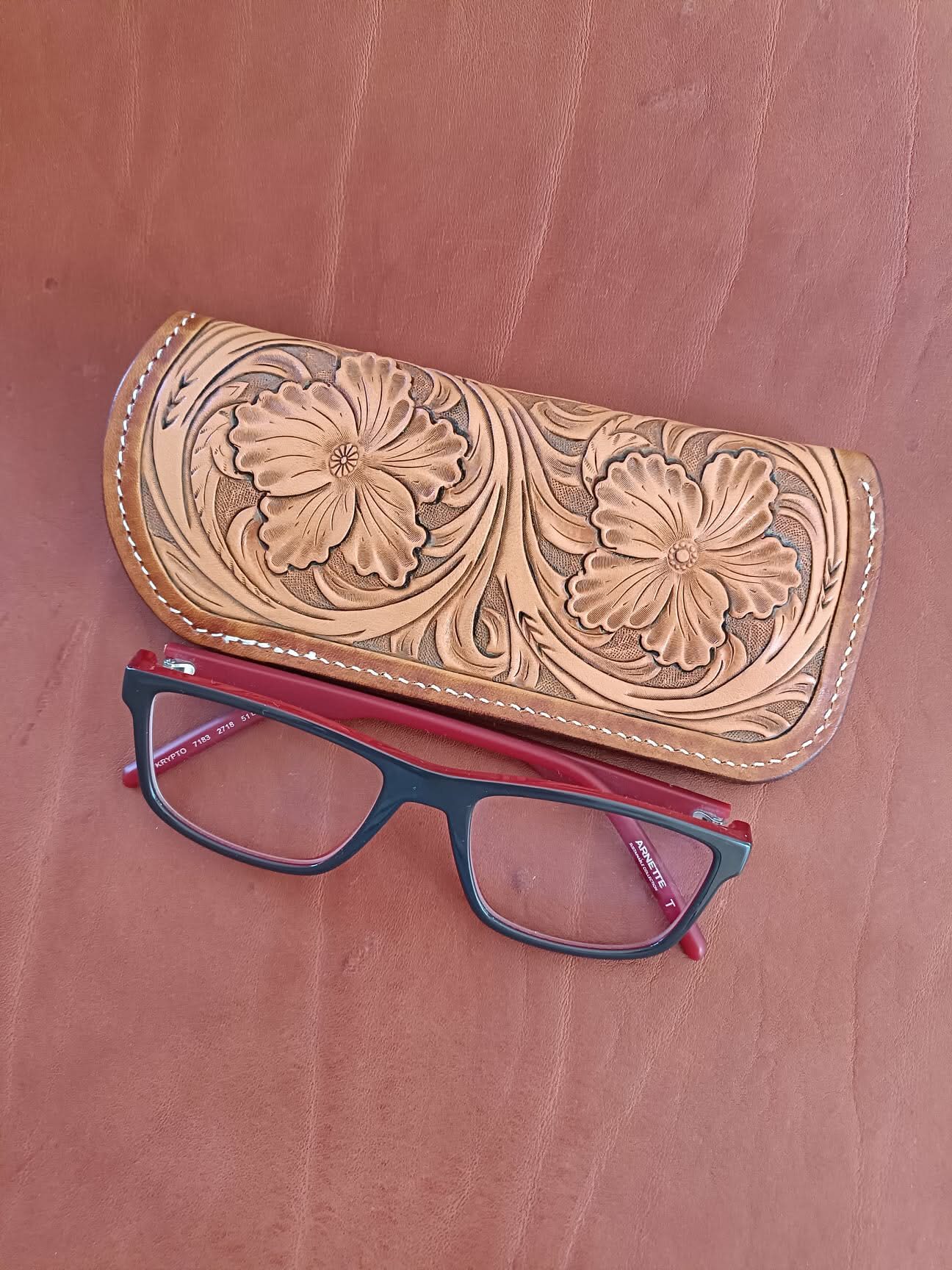 Étui à lunettes en cuir naturel avec un grand motif de fleurs sculptées en relief et coutures blanches, posé à côté d'une paire de lunettes sur un fond en cuir marron.