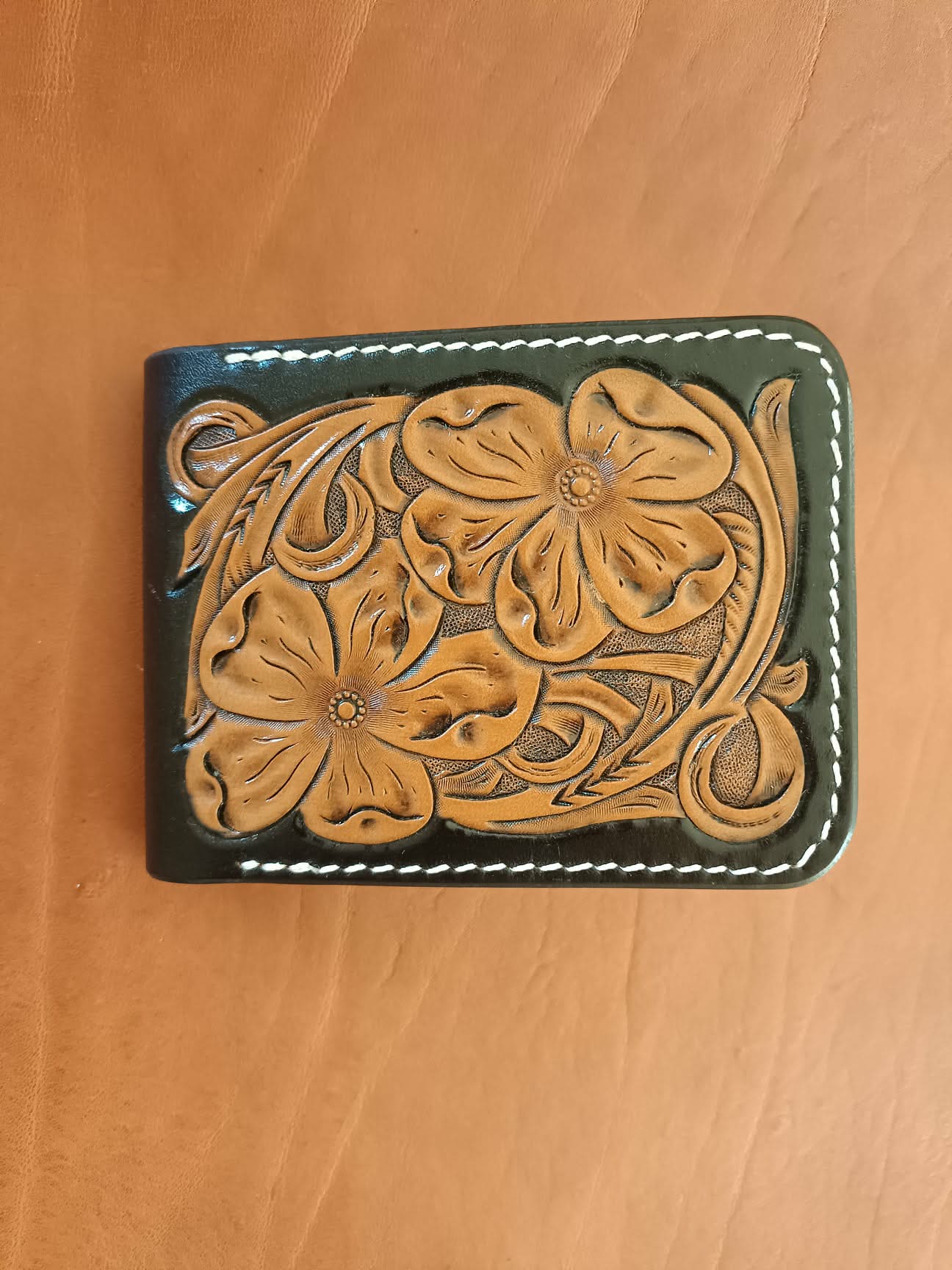 Face avant d'un portefeuille bifold en cuir noir avec motif floral brun repoussé à la main et coutures blanches.