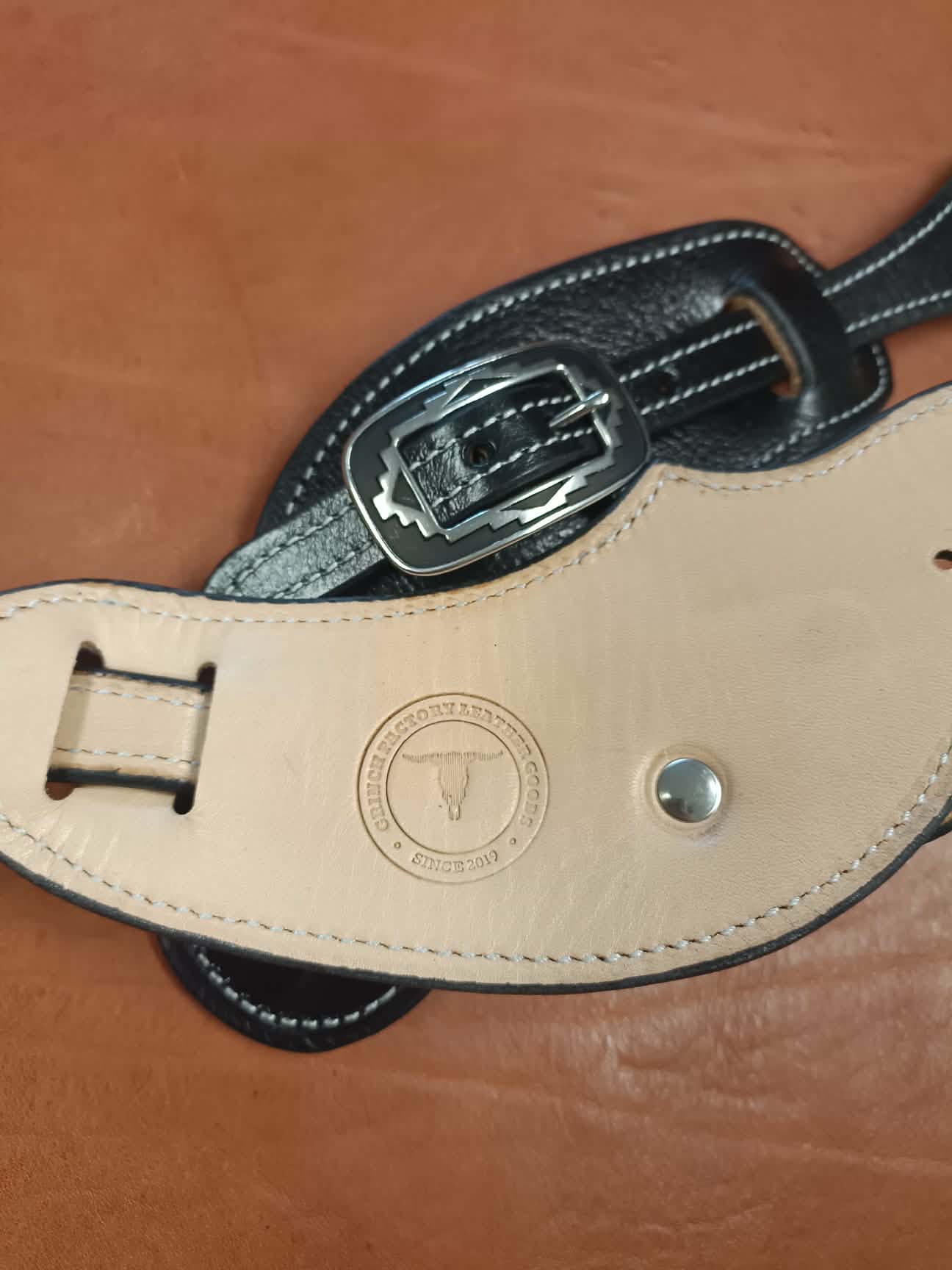 Paire de pattes d'éperons en cuir noir artisanal avec coutures blanches et boucles métalliques gravées style Western posées sur un fond en cuir marron.