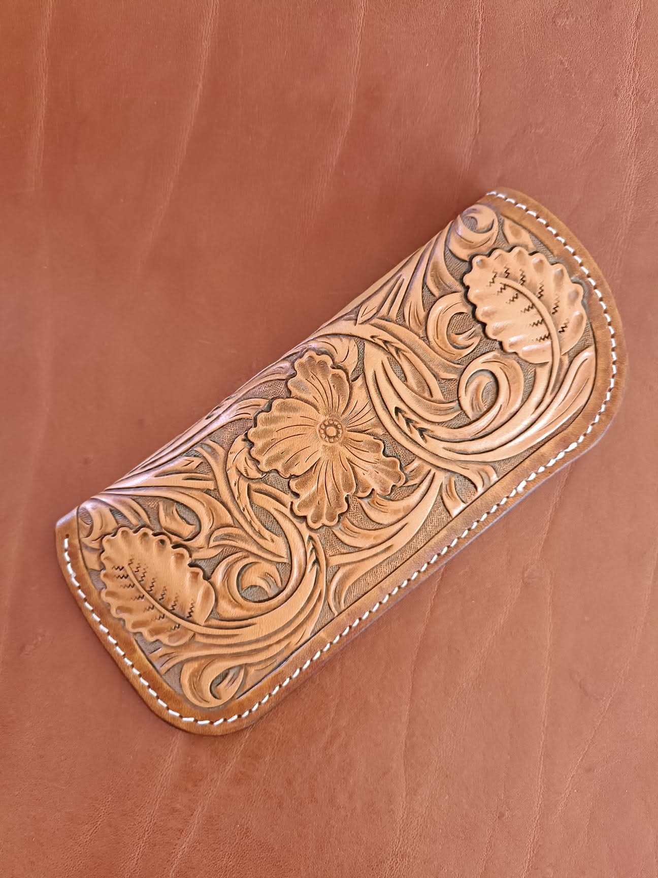 Étui à lunettes en cuir naturel avec un grand motif de fleurs sculptées en relief et coutures blanches, posé à côté d'une paire de lunettes sur un fond en cuir marron.