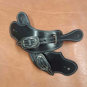 Paire de pattes d'éperons en cuir noir artisanal avec coutures blanches et boucles métalliques gravées style Western posées sur un fond en cuir marron.
