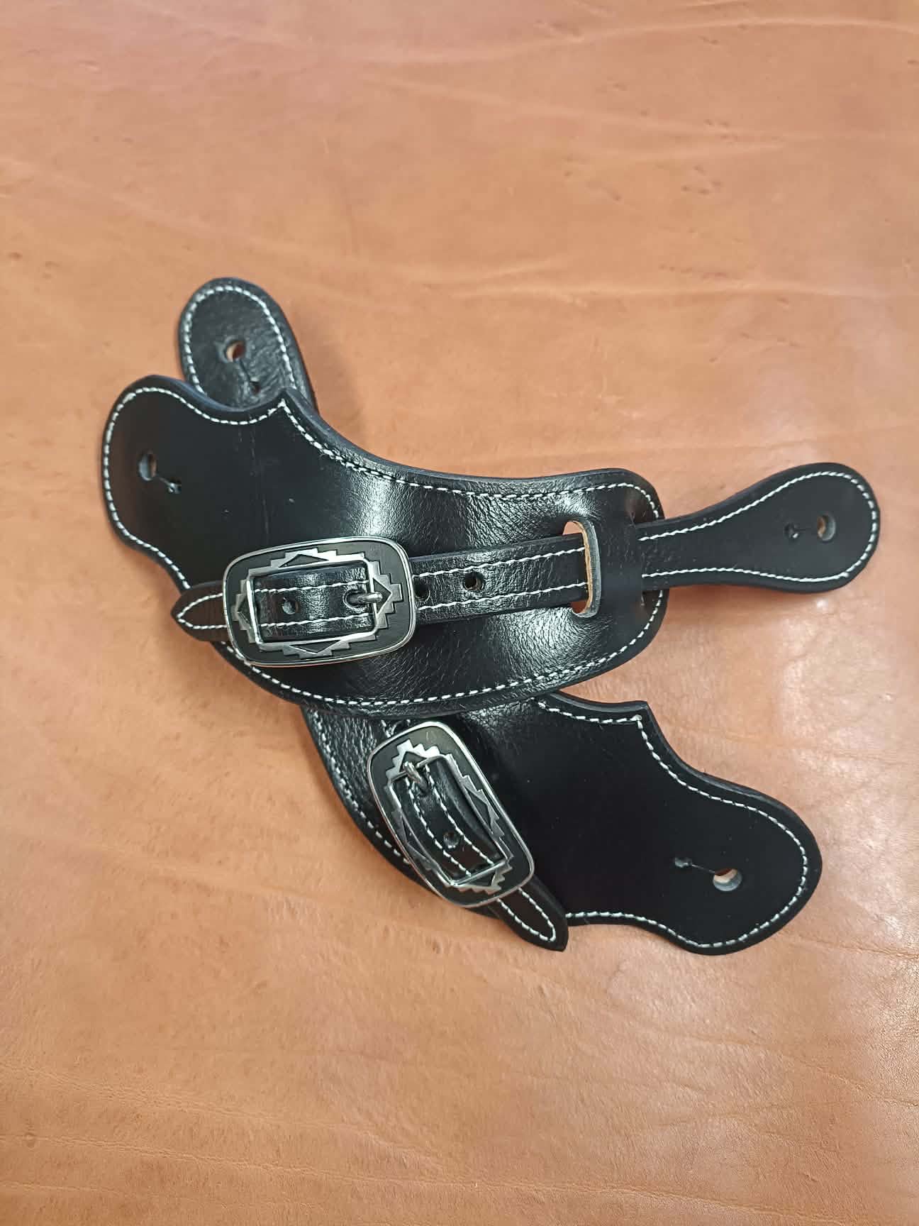 Paire de pattes d'éperons en cuir noir artisanal avec coutures blanches et boucles métalliques gravées style Western posées sur un fond en cuir marron.
