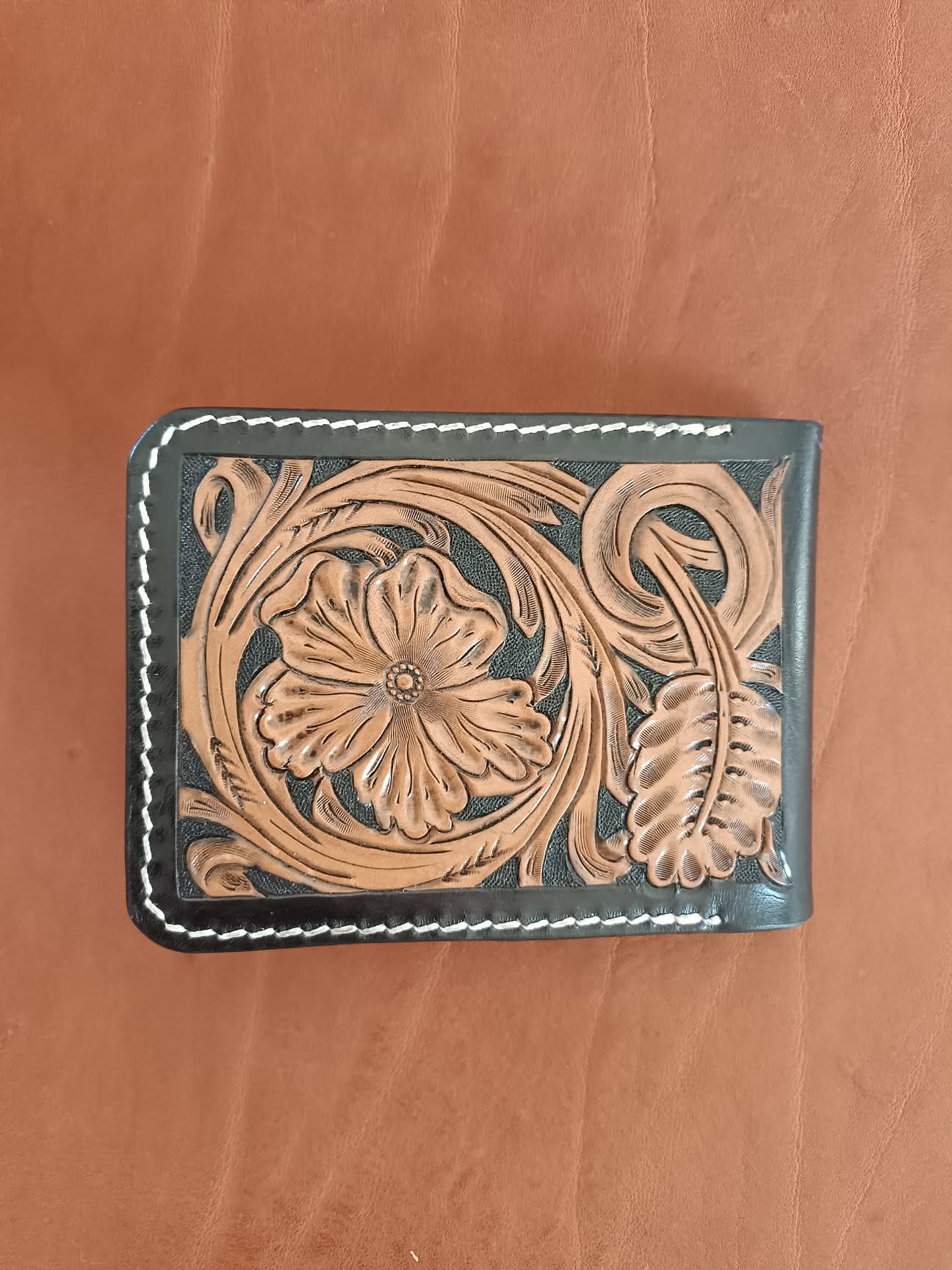 Face avant d'un portefeuille bifold en cuir noir avec motif floral brun repoussé à la main et coutures blanches.