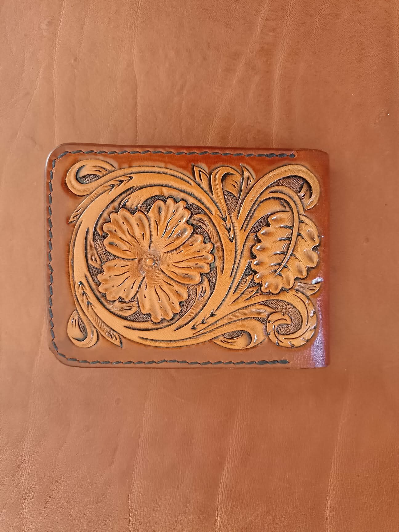 Face avant d'un portefeuille bifold en cuir fauve avec motif floral brun repoussé à la main et coutures noires.
