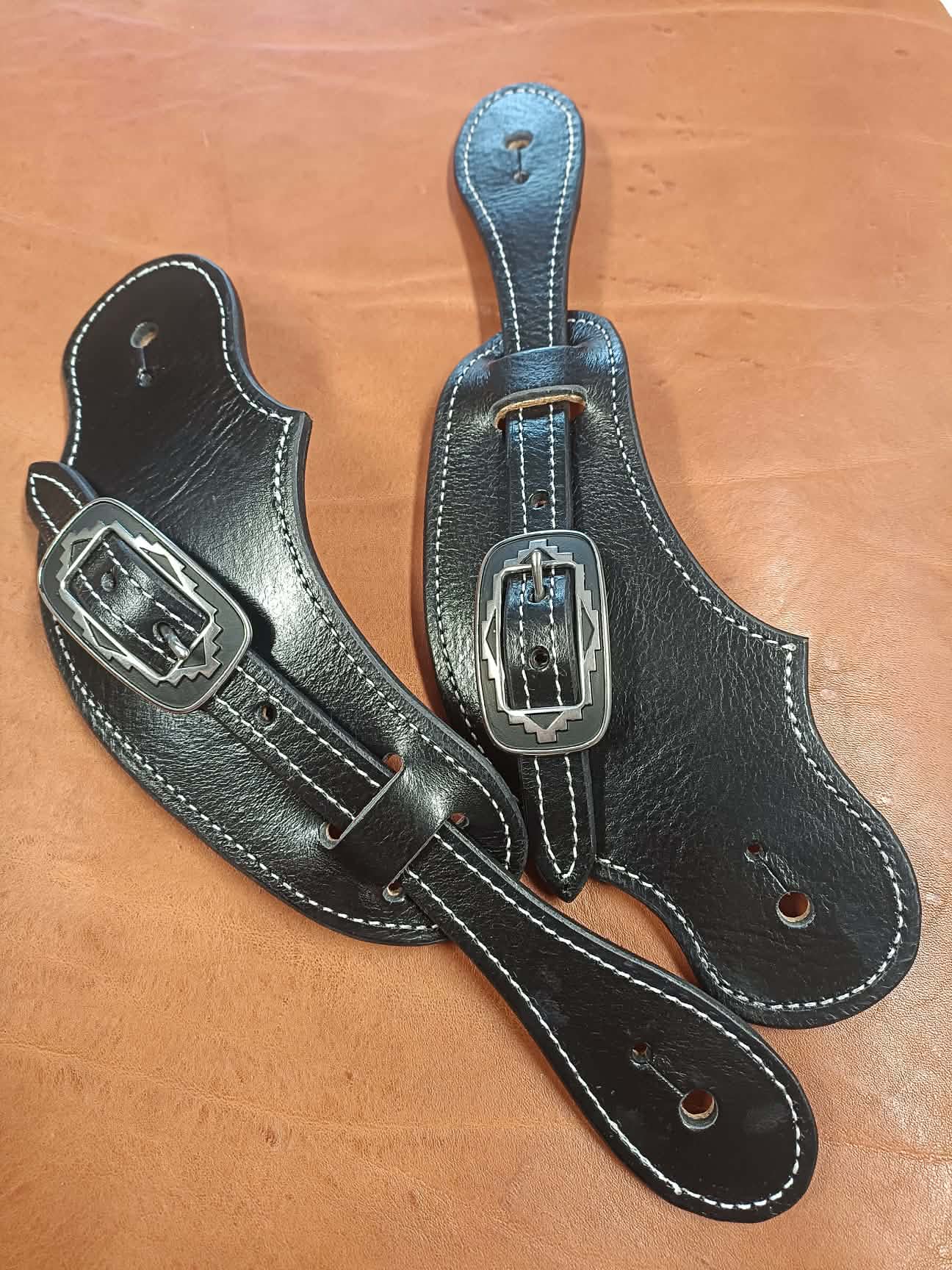 Paire de pattes d'éperons en cuir noir artisanal avec coutures blanches et boucles métalliques gravées style Western posées sur un fond en cuir marron.