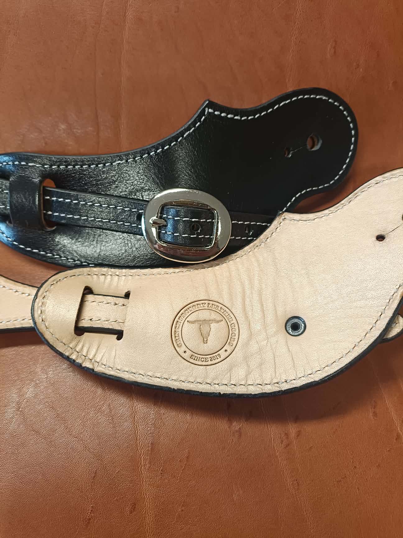 Paire de pattes d'éperons en cuir noir artisanal avec coutures blanches et boucles lisses posées sur un fond en cuir noir.