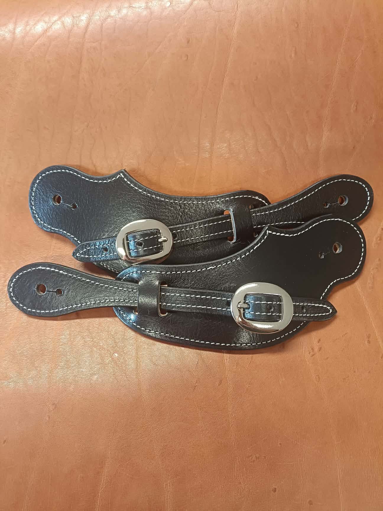 Paire de pattes d'éperons en cuir noir artisanal avec coutures blanches et boucles lisses posées sur un fond en cuir noir.